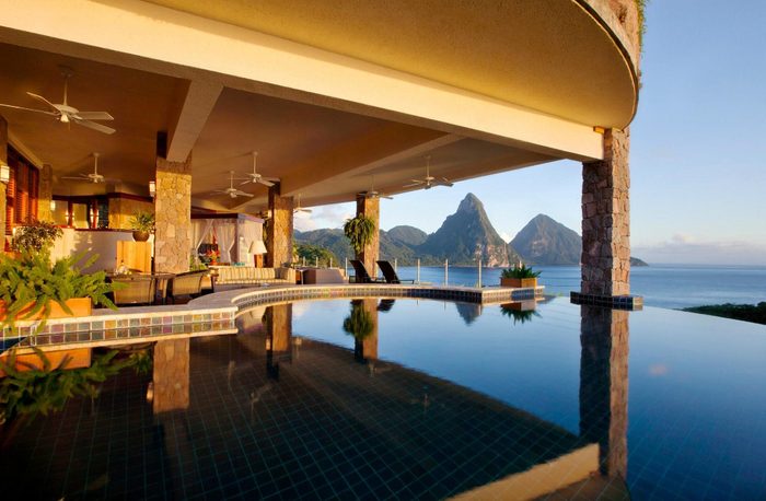 Infinity pool reflects mountains, under a stone-pillared terrace, with lounge chairs and plants, overlooking a calm sea at sunset.