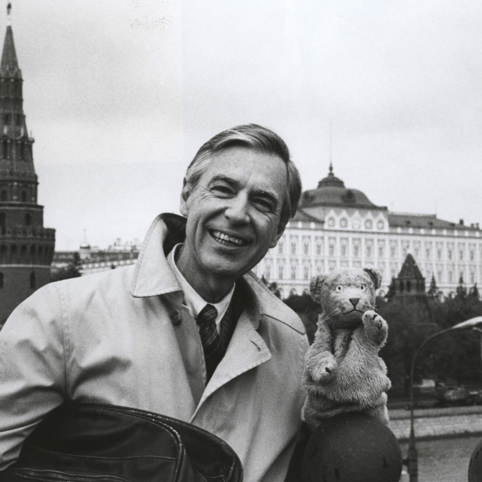 Man smiling, holding a puppet, stands near a river with the Kremlin's towers and buildings prominently in the background.