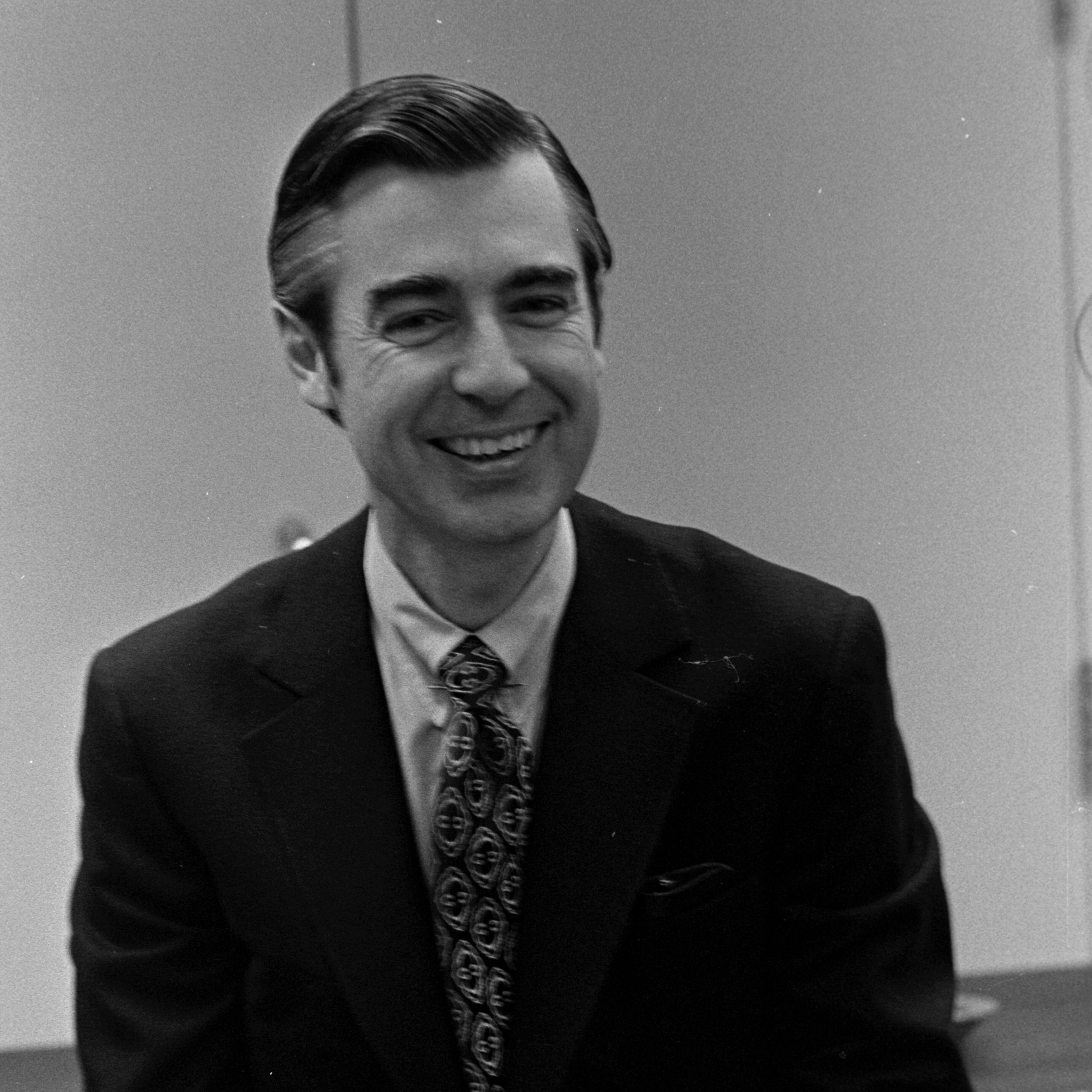 Man smiling, hands outstretched, wearing a suit and patterned tie; sitting indoors, against plain walls and a partial view of a table.