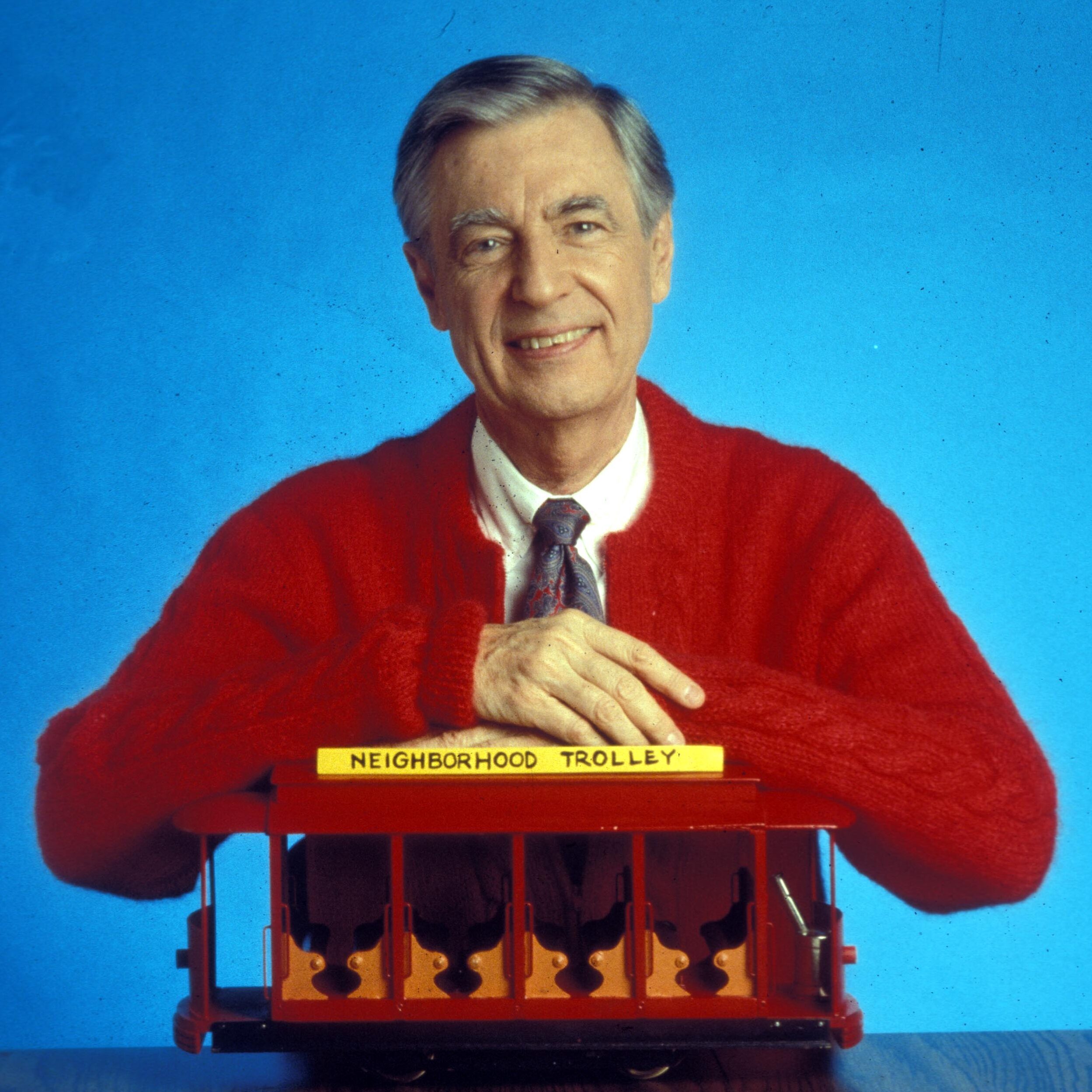 Man sitting, smiling beside a small red trolley labeled "NEIGHBORHOOD TROLLEY," wearing a red sweater; blue background.