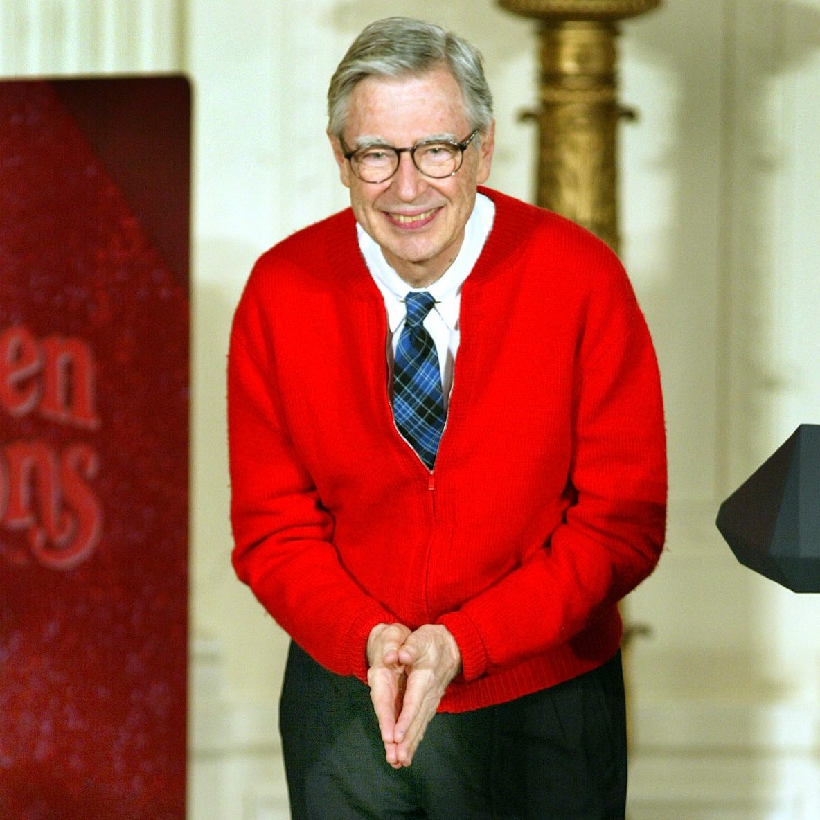 An elderly man wearing a red sweater smiles and bows slightly indoors near a podium.