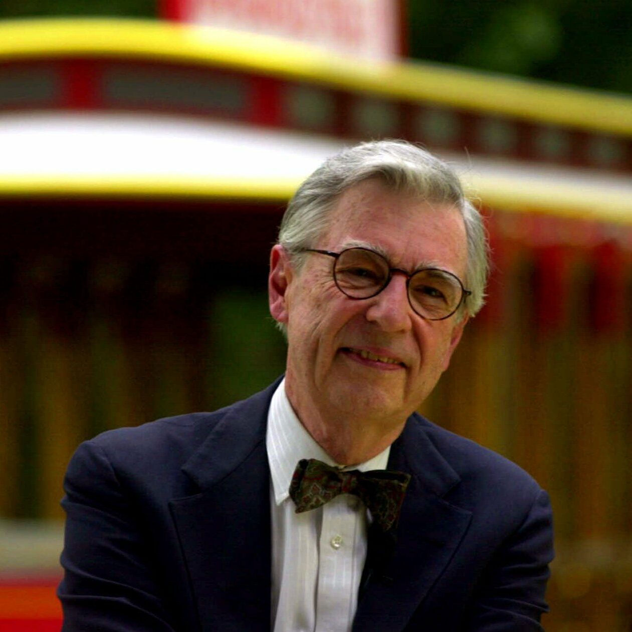 Man smiles, wearing glasses and bow tie, standing outside near a colorful trolley.