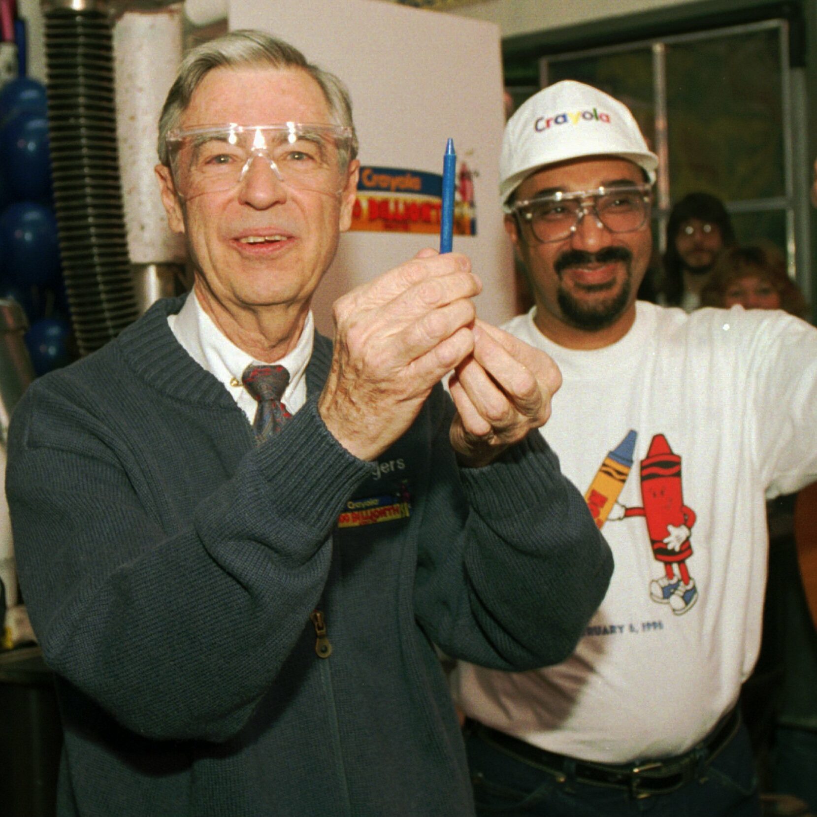 Man holds a blue crayon, wearing goggles indoors; another man in a Crayola hat gives a thumbs-up.