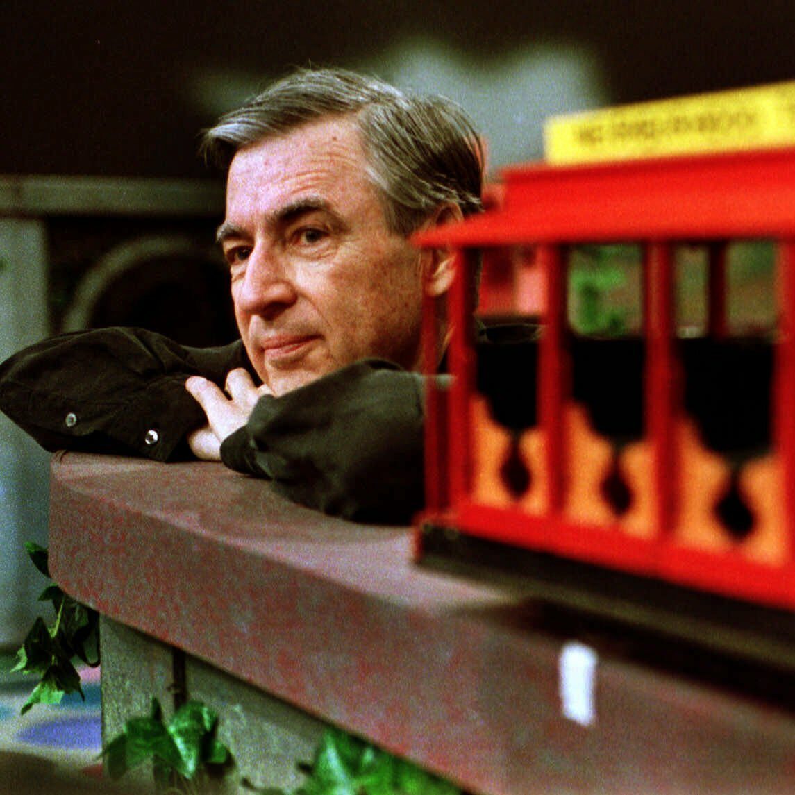 A man gazes thoughtfully beside a red toy trolley car on a set with green foliage.