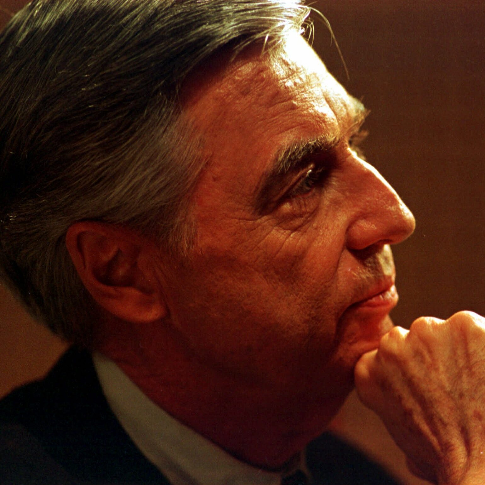 A person with gray hair looks thoughtful, resting their chin on their hand, in a softly lit environment.
