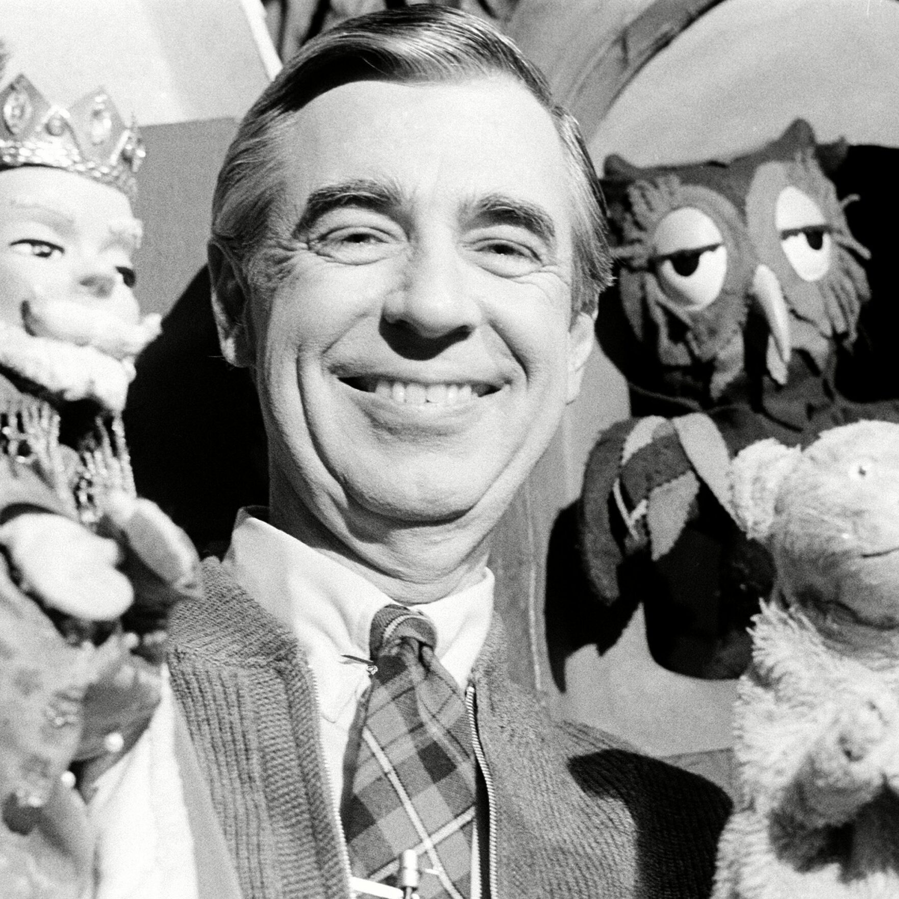 Man smiling while holding puppets; background includes additional puppets. The setting suggests a friendly and playful environment.