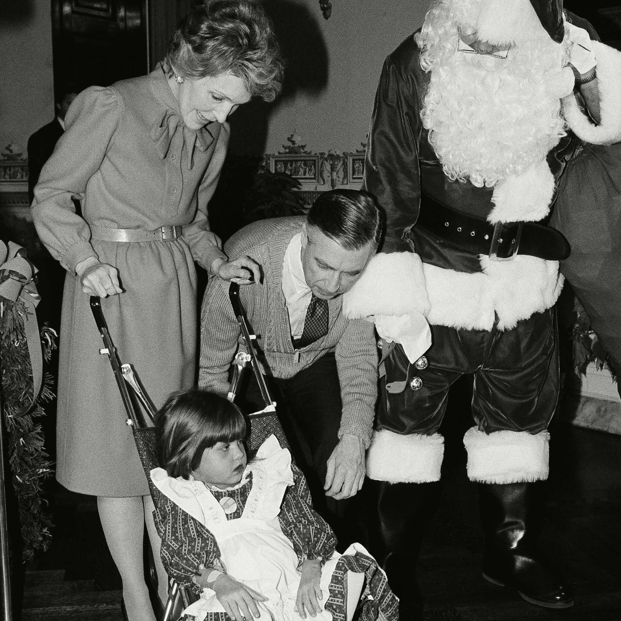 A child sits in a stroller wearing a dress, surrounded by standing adults, one dressed as Santa, in an indoor setting.