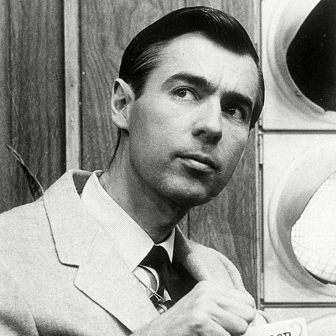 Man holding a sign with text, "WON'T YOU BE MY NEIGHBOR MISTER ROGERS," indoors, against a wooden background with a circular object nearby.