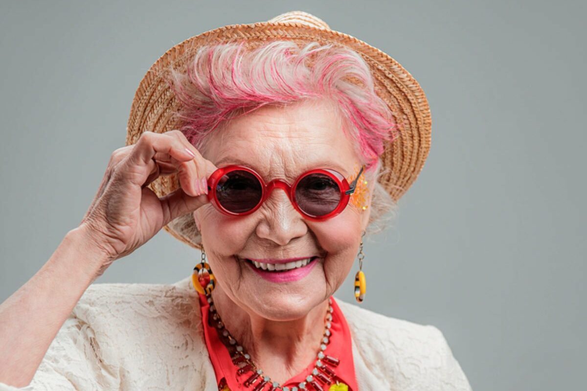 A smiling elderly woman adjusts red sunglasses, wearing a straw hat and colorful jewelry, standing against a plain background.