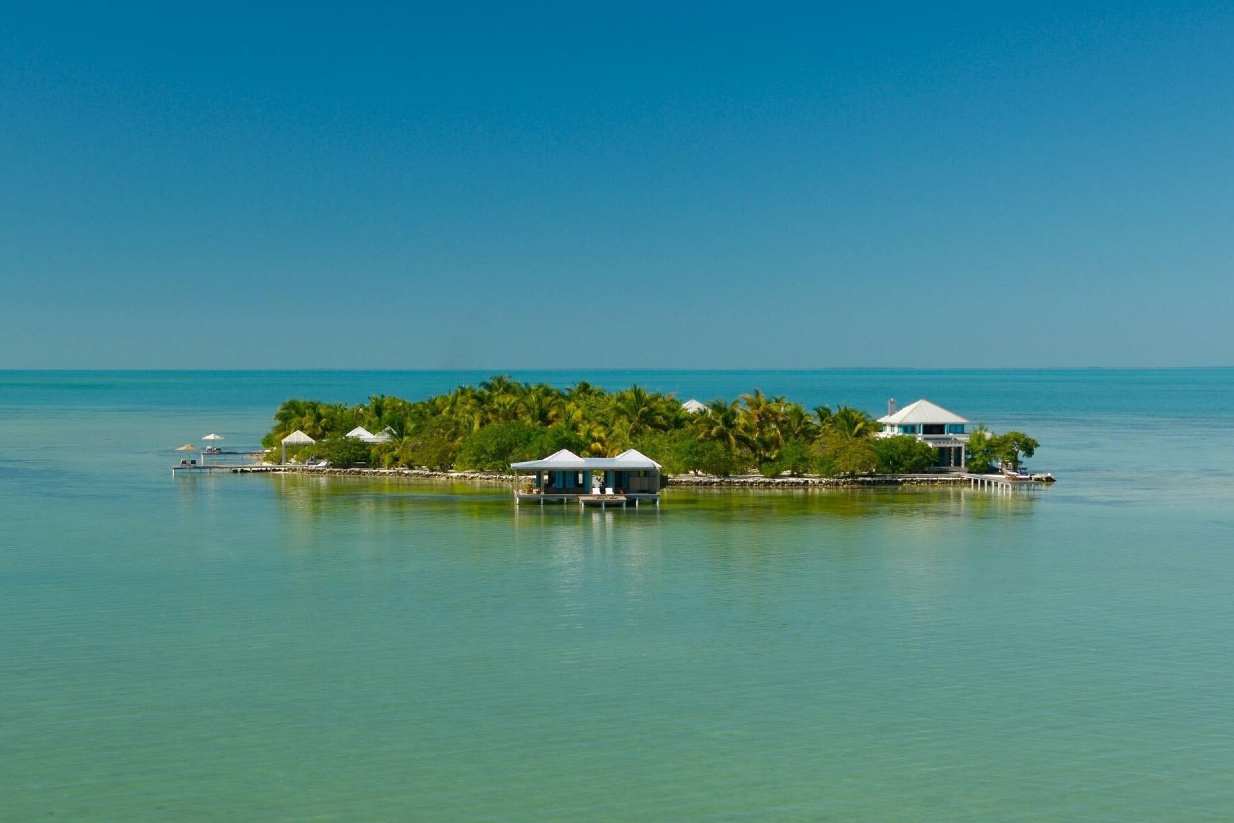 Small island with palm trees and houses rests in calm turquoise sea under a clear blue sky.
