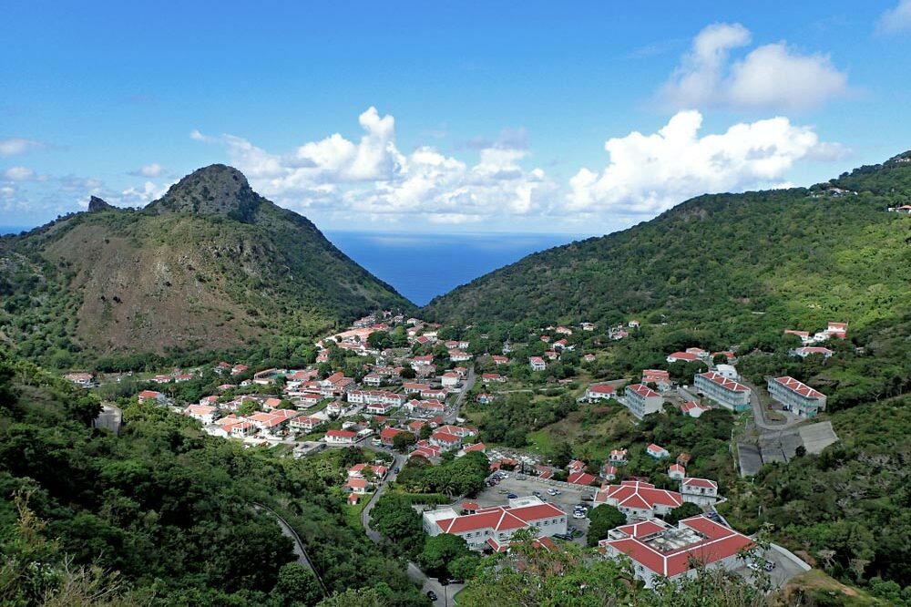 A cluster of red-roofed buildings nestles in a lush, hilly landscape, with a mountain peak and ocean visible in the background.