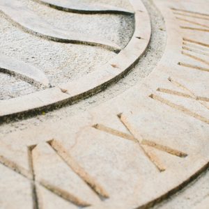 Stone sundial displays Roman numerals in circular sequence, positioned outdoors in a rustic setting.