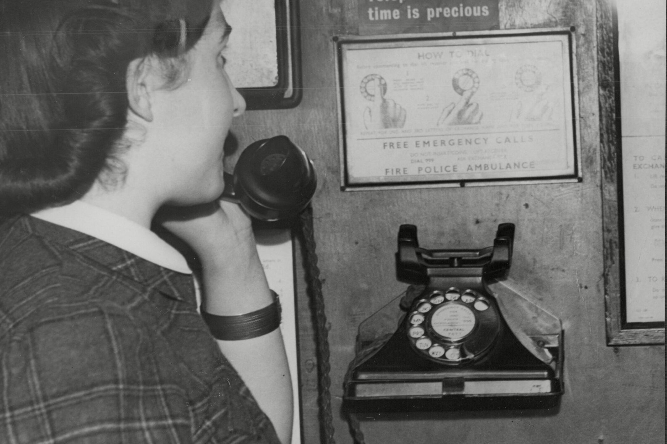 Woman uses a rotary phone in a wooden booth, surrounded by instructions and a sign reading, "Keep your talks short! Telephone line-time is precious."