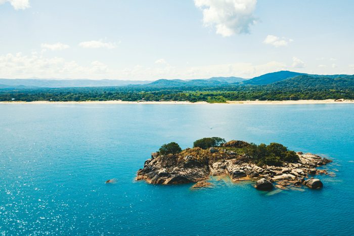 A small rocky island with greenery sits in calm blue water, backed by a distant shoreline and forested hills under a partly cloudy sky.