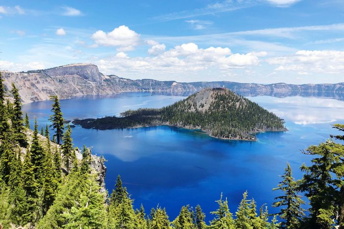 An island sits surrounded by deep blue water, encircled by pine-covered hills beneath a clear sky with scattered clouds.