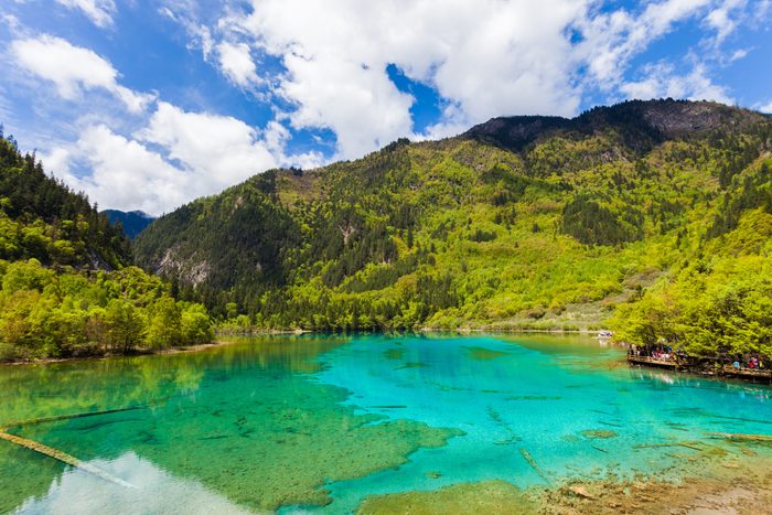 Clear turquoise lake reflects lush green mountains under a bright blue sky with scattered white clouds.