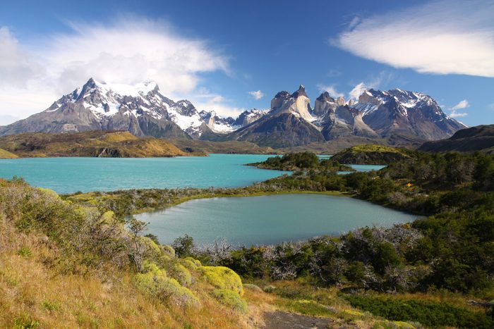 Mountains rise majestically behind turquoise lakes surrounded by lush green foliage, under a clear blue sky with scattered clouds.