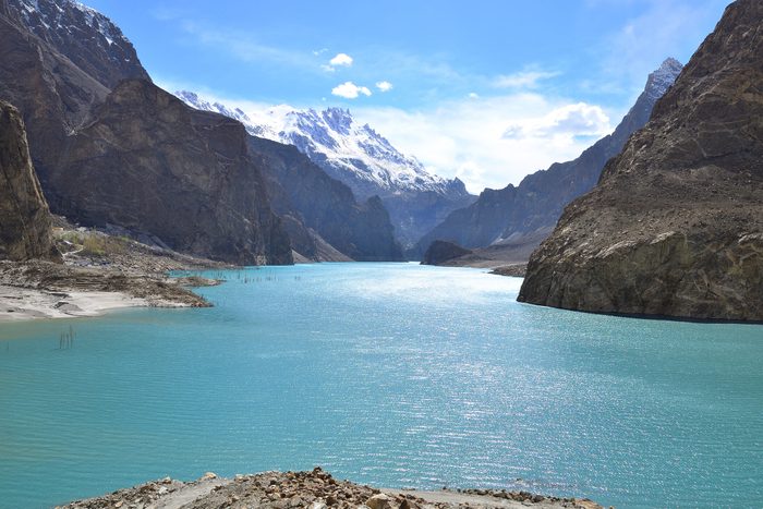 A turquoise river flows between towering rocky mountains under a bright blue sky with scattered white clouds.