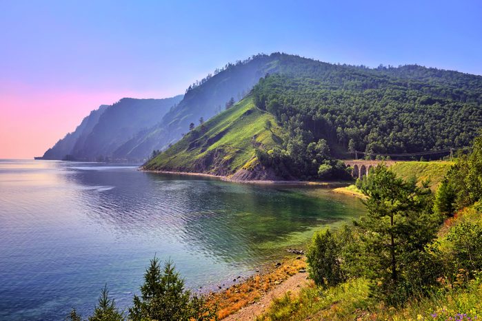 Mountain meets a calm lake, surrounded by lush greenery and vibrant flowers, under a clear blue sky at sunrise or sunset.