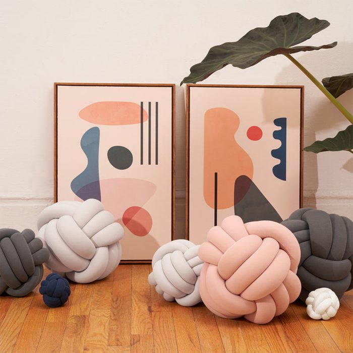 Knot pillows rest on wooden floor; abstract art frames lean against wall; large leafy plant provides decor context.