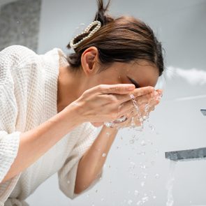 Person washing face with hands cupped under flowing faucet in a bathroom, wearing a white robe, with hair held by a clip.