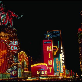 Neon signs illuminate, advertising casinos and hotels at night, with colorful lights and a large cowgirl figure atop "Golden Goose."