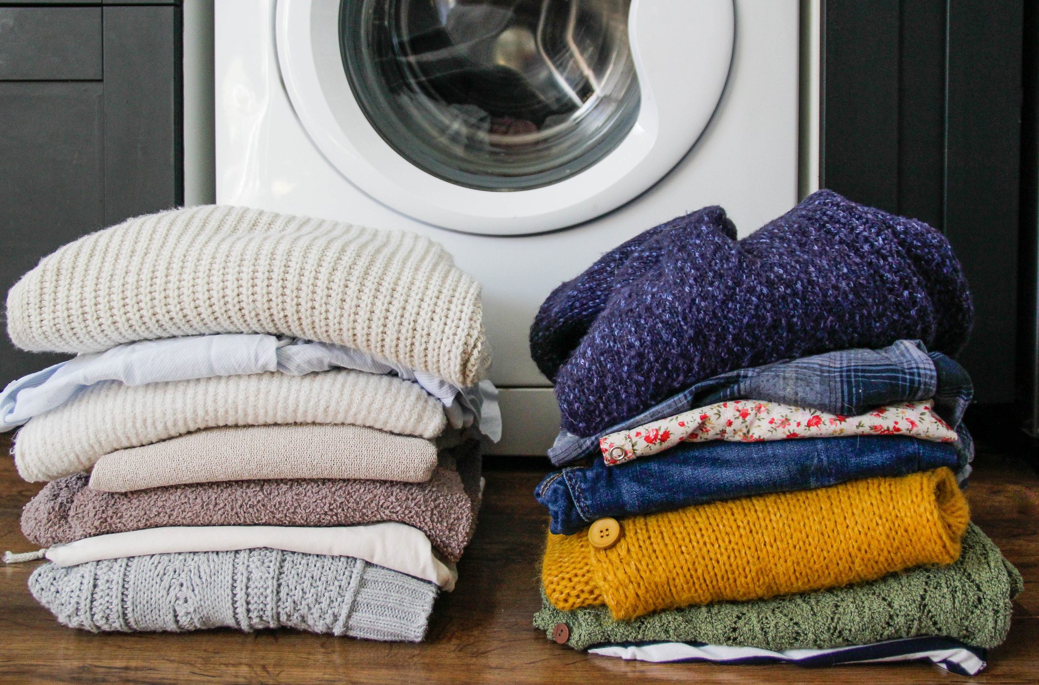 Two stacks of folded sweaters rest on a wooden floor in front of a white washing machine.