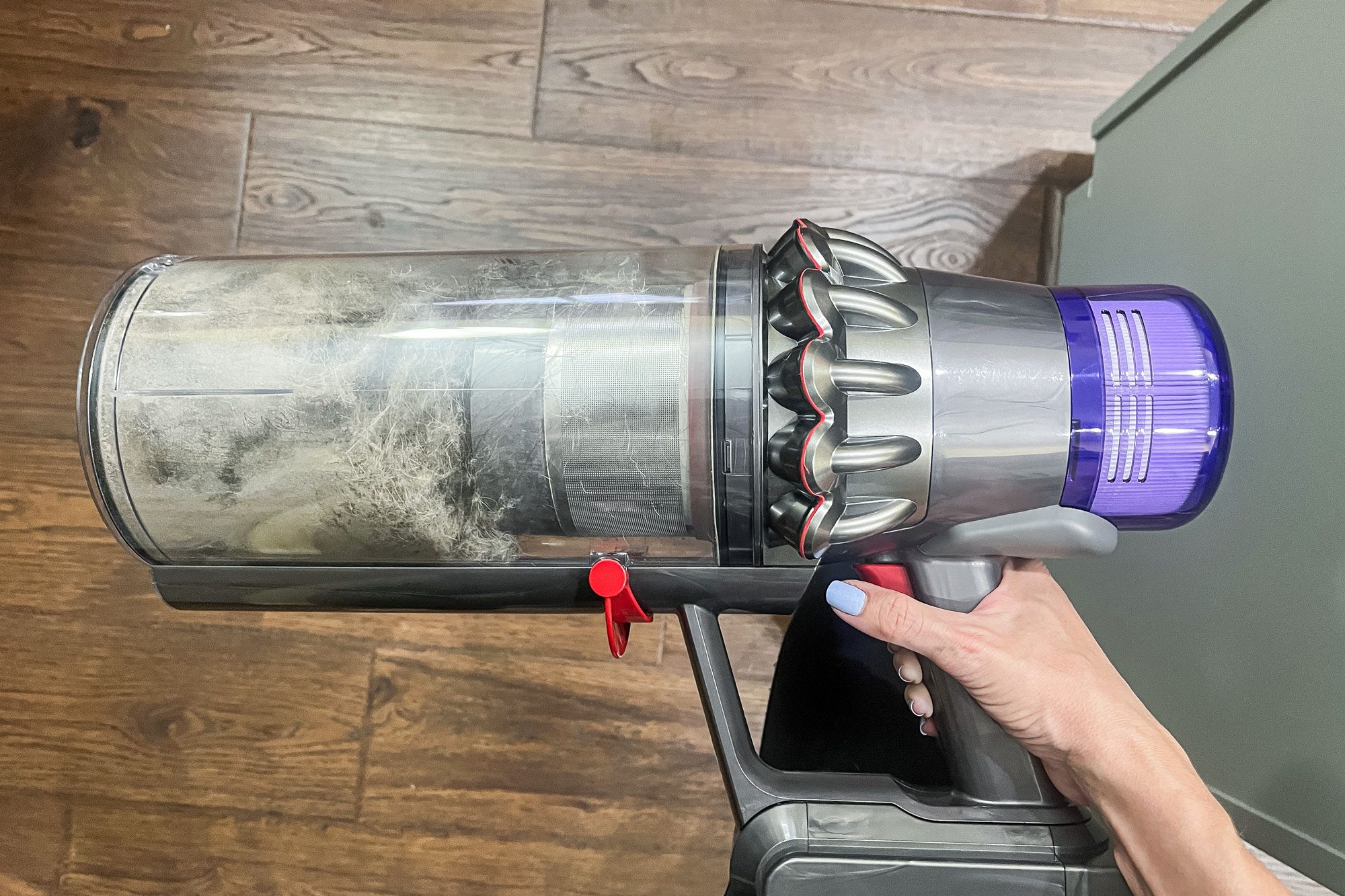 Hand holds a vacuum cleaner filled with dust. The setting is a wooden floor, and the vacuum is gray and purple.