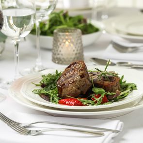 Steak sits on arugula with red peppers, accompanied by glasses of red and white wine on a white tablecloth with elegant table setting.