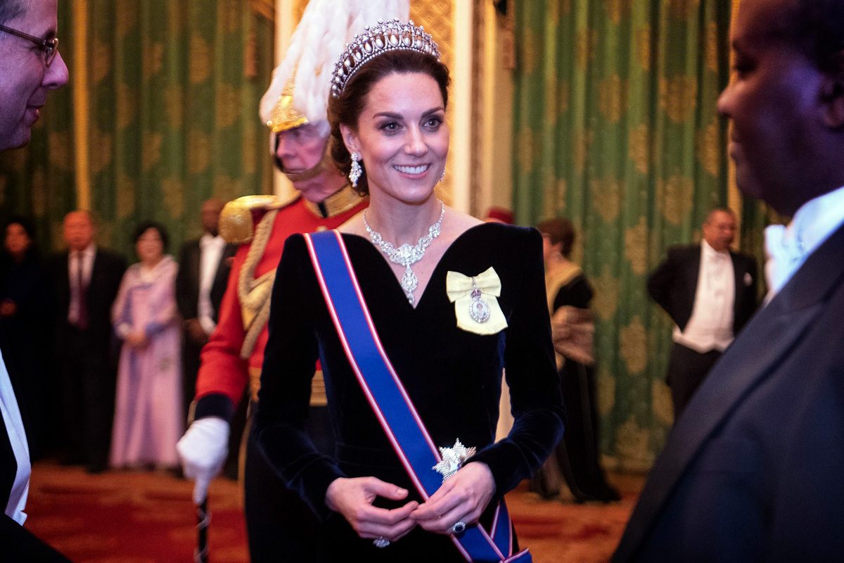 Kate Middleton wearing a tiara smiles in formal attire, engaging with people at a grand indoor event with elaborately decorated walls.