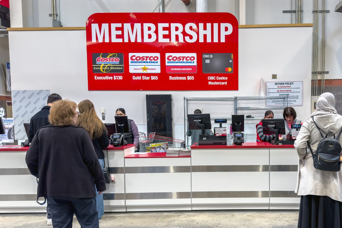 A Costco membership counter where customers are being assisted by employees, with a large sign displaying membership options