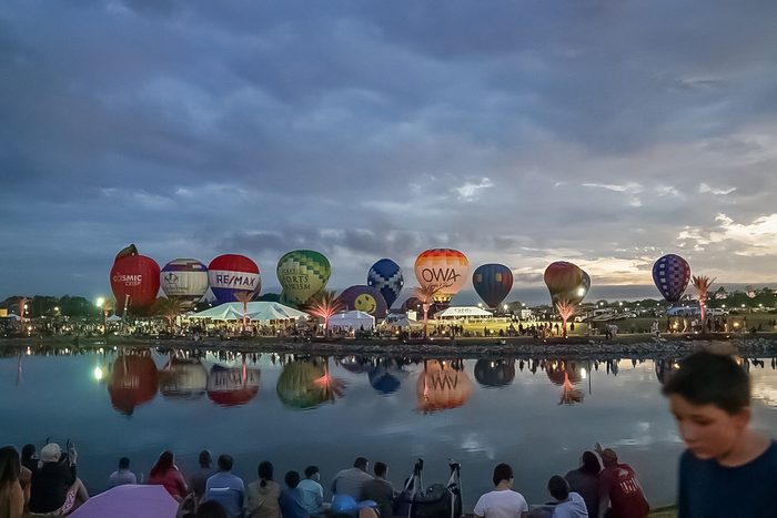 A row of colorful hot air balloons is lined up along the edge of a calm lake at dusk, reflecting in the water, with many people gathered on the shore watching the event under a cloudy sky.